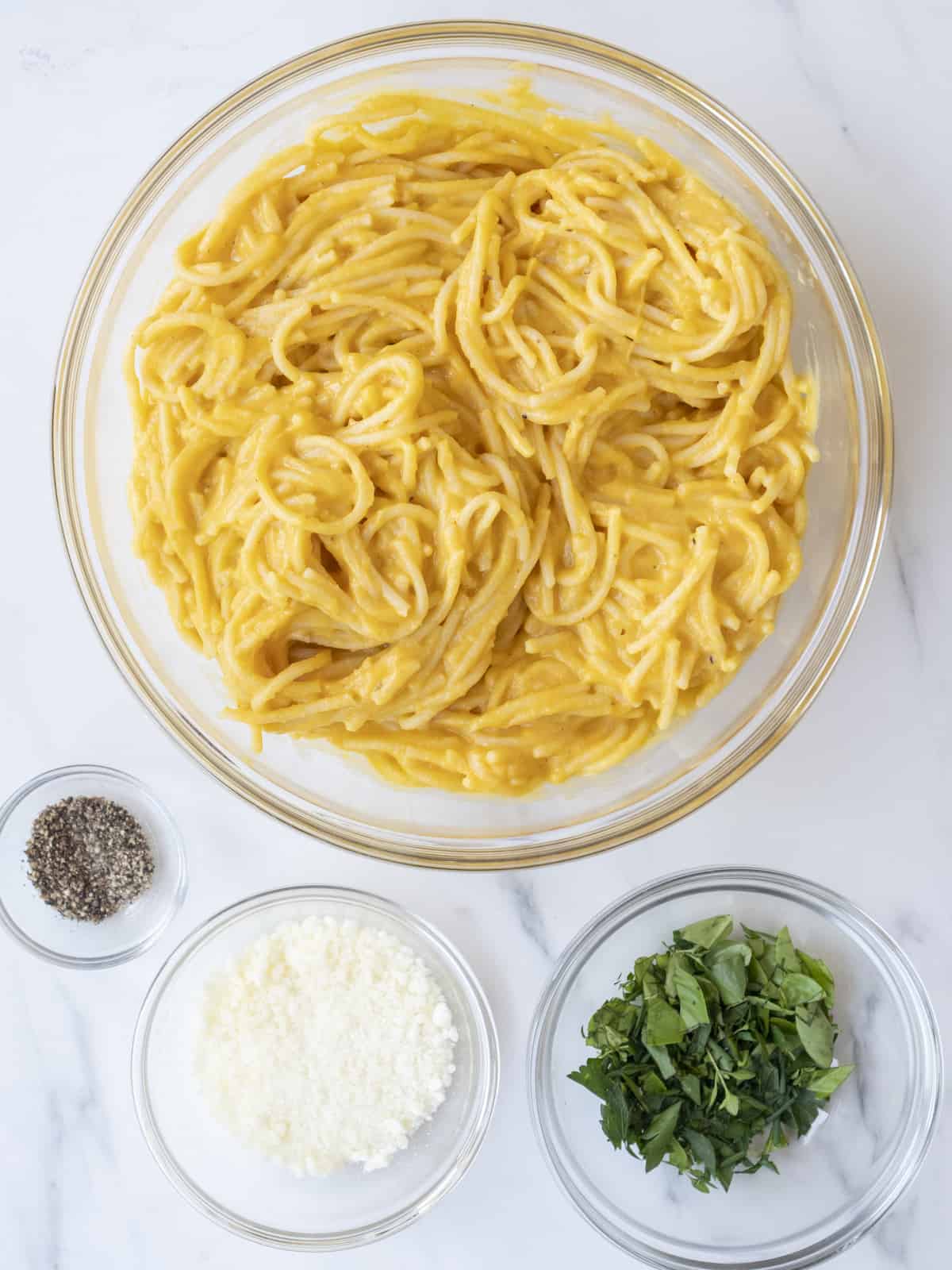 Butternut squash pasta in a glass bowl next to a serving bowl of pepper, a bowl of pecorino, and a bowl of fresh herbs.  