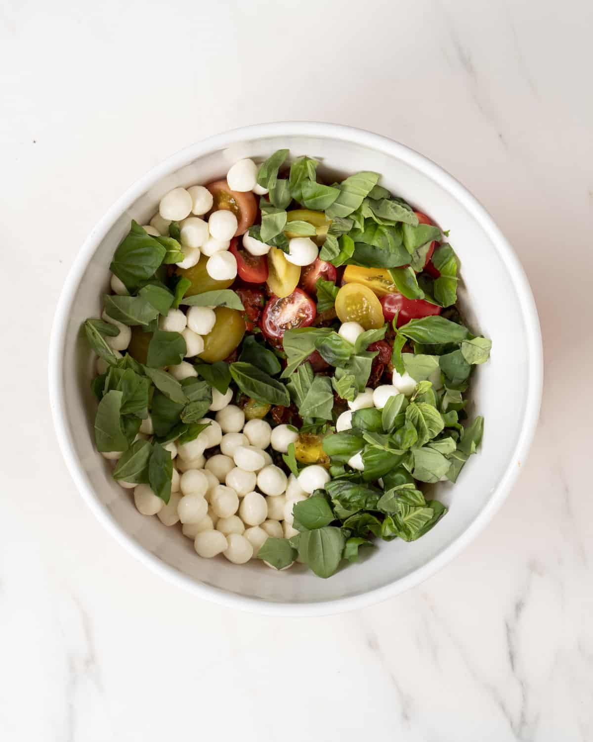 Large white mixing bowl with orzo topped with lots of fresh cherry tomatoes, mozzarella pearls and basil.