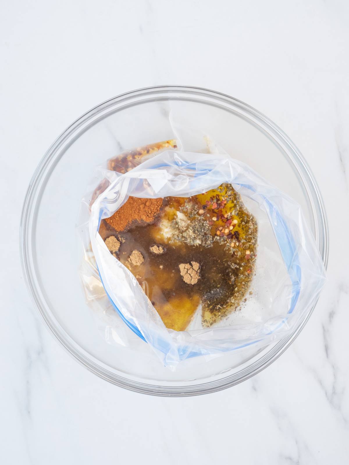A glass mixing bowl with a zip-top bag with all the seasonings along with lemon juice and olive oil to make chicken marinade.