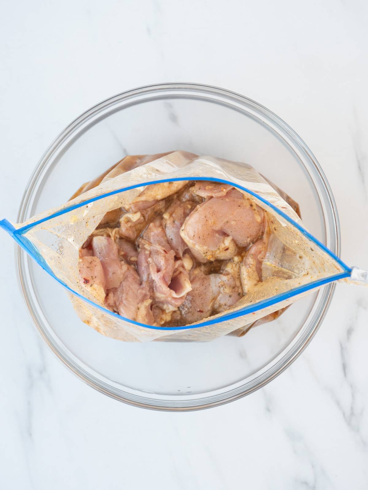 A glass mixing bowl with a zip-top bag with the chicken marinated in a spices and olive oil marinade.