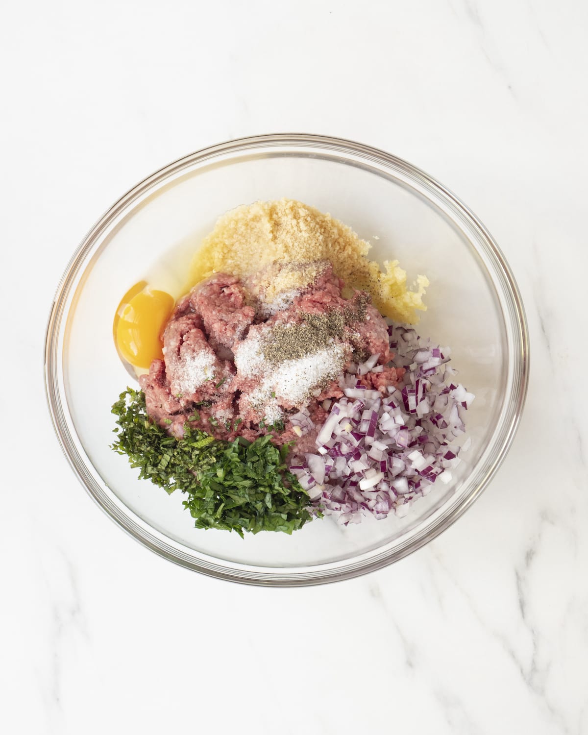 A clear bowl with ground lamb, parsley, mint, red onion, garlic, breadcrumbs, egg, salt and pepper.