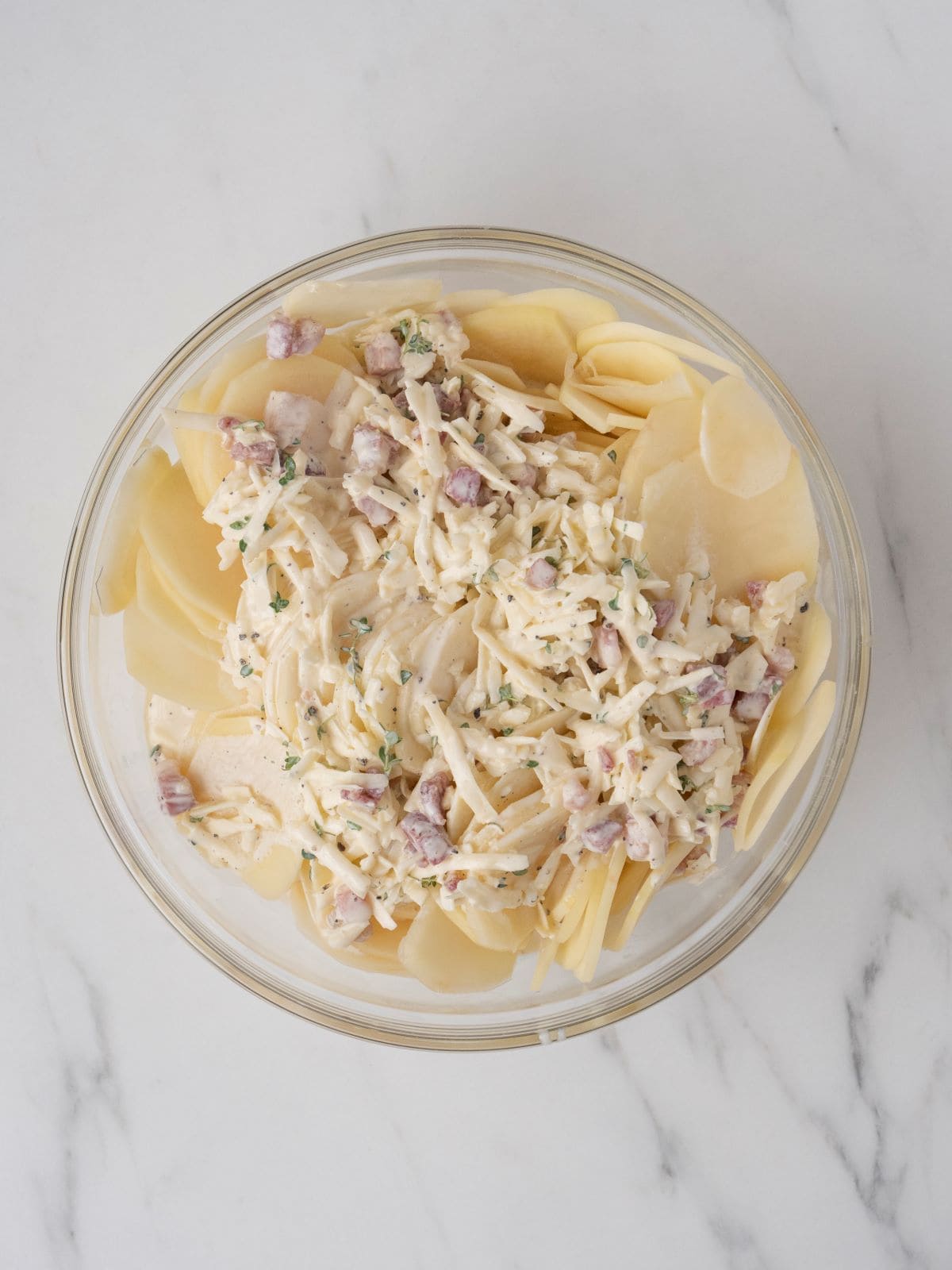 A big glass mixing bowl with sliced potatoes along with the cream mixture.