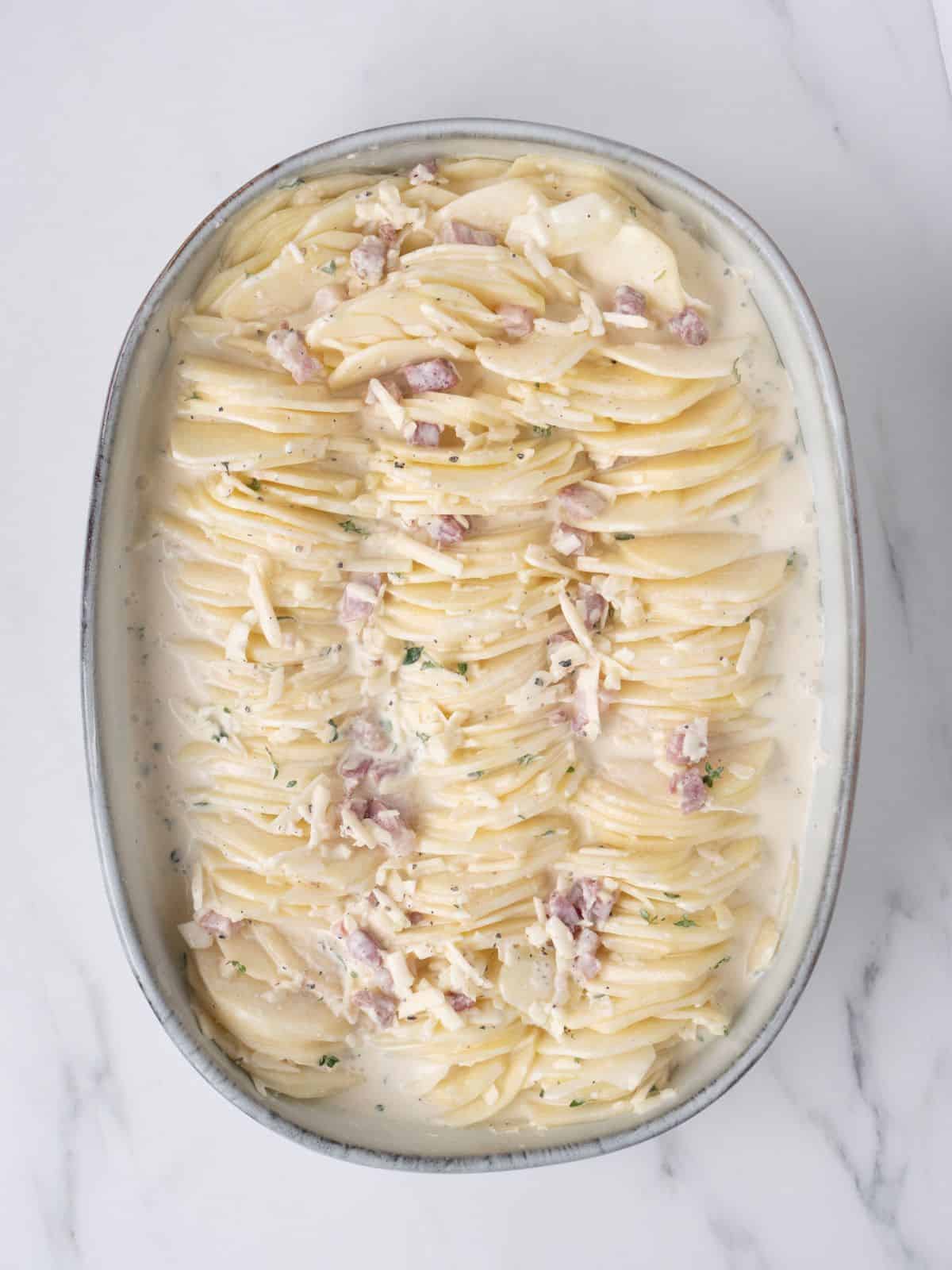 An oval baking dish with sliced potatoes dipped in cream mixture arranged in three columns along with the leftover cream mixture poured on top.