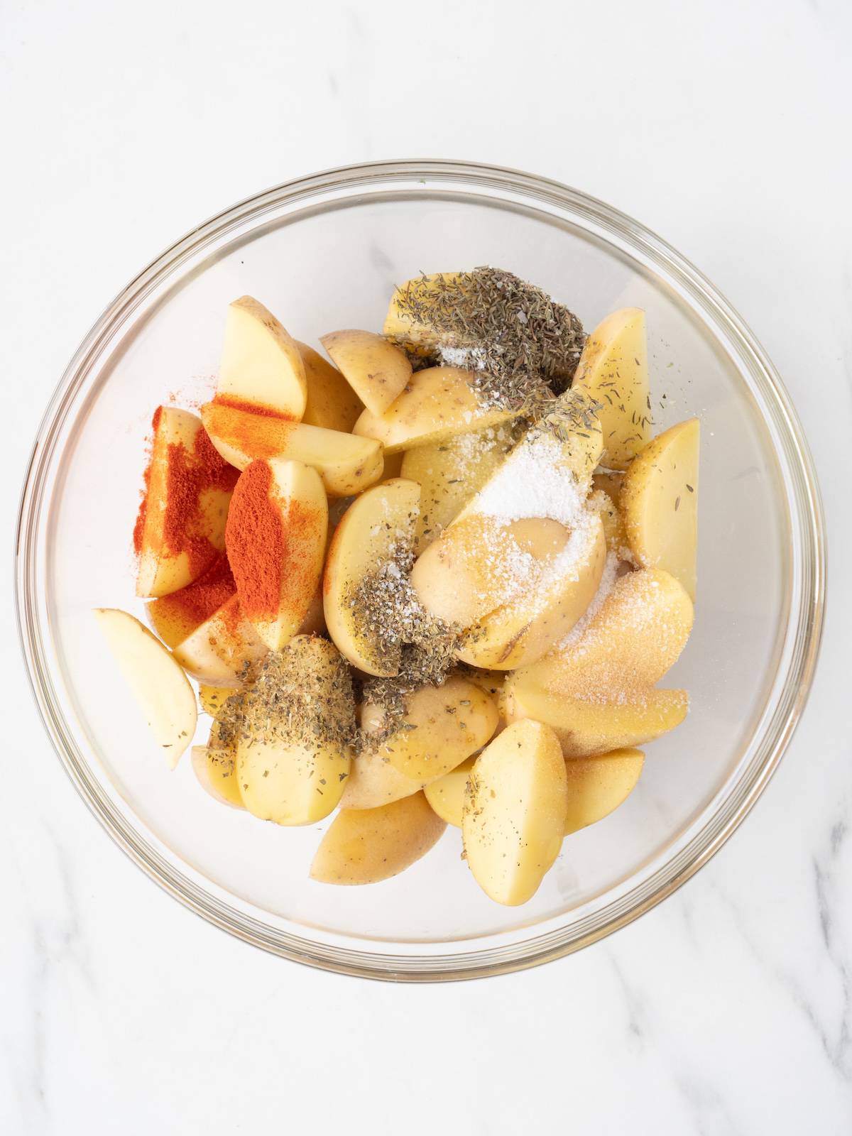 A large glass mixing bowl with quartered potatoes, seasonings and olive oil.
