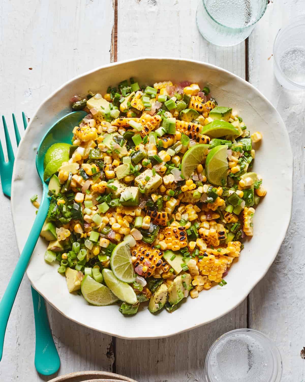 A white bowl with grill corn and avocado salad, garnished with lime wedges and herbs and a fork and spoon.