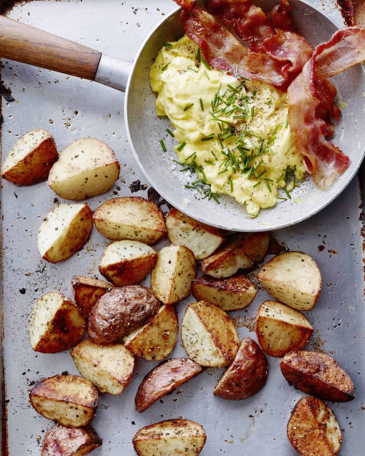 A skillet with eggs and bacon topped with chives sitting next to a baking sheet with cooked potato wedges.