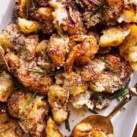 An overhead shot of cubed golden crispy roasted potatoes with melted parmesan cheese and chives on a white rectangular serving platter with a gold serving spoon on a mustard yellow linen napkin on a wooden table