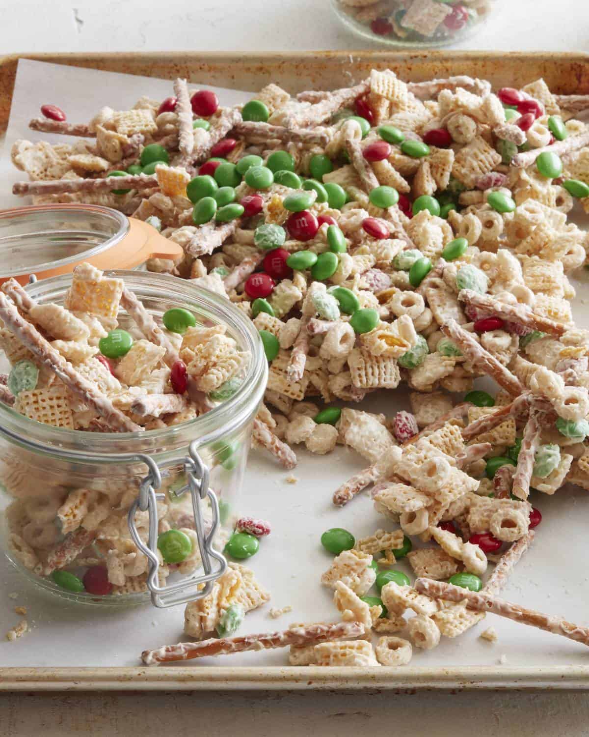 A parchment-lined baking sheet with christmas mix and an open glass canister with more christmas mix in it.