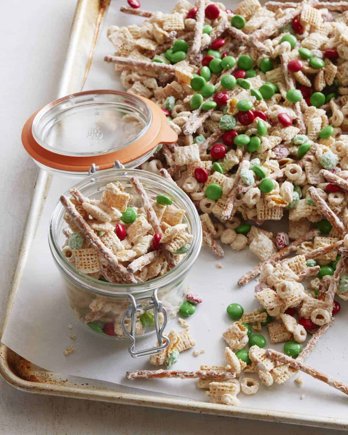 A parchment-lined baking sheet with christmas mix and an open glass canister with more christmas mix in it.