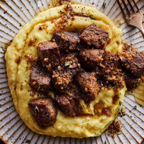 An overhead shot of a ceramic fluted plate with garlic butter steak bites over creamy polenta. There is a fork resting on the right rim of the plate and a burn orange linen napkin to the left side of the plate.