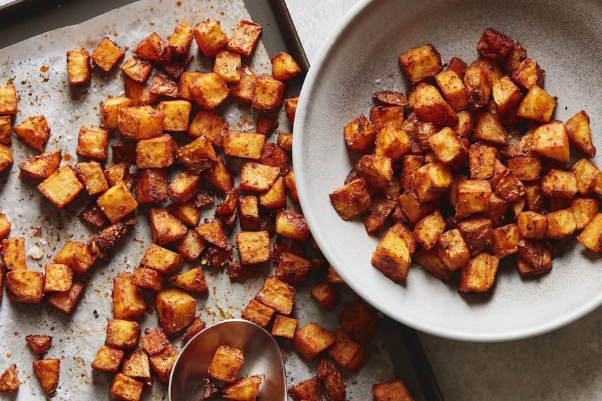 A close-up shot of a parchment paper-lined baking sheet with cubed roasted potatoes and a serving spoon, and a grey bowl on the side with more potatoes in it.