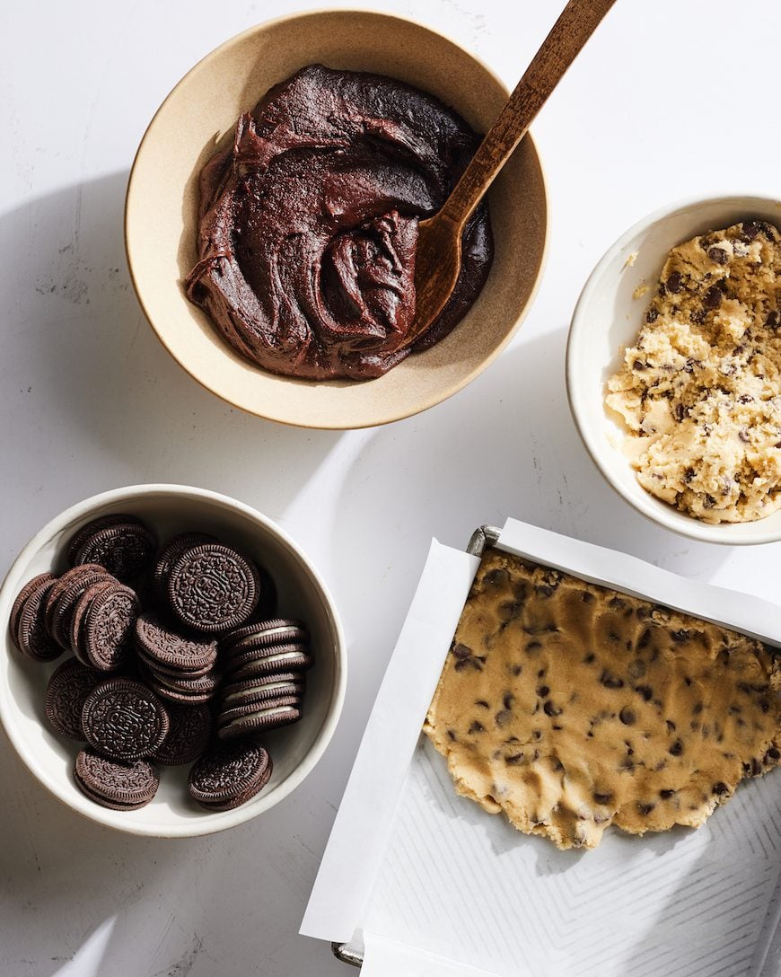 A bowl of oreos, brownie batter, cookie dough and a baking pan with cookie dough in it.
