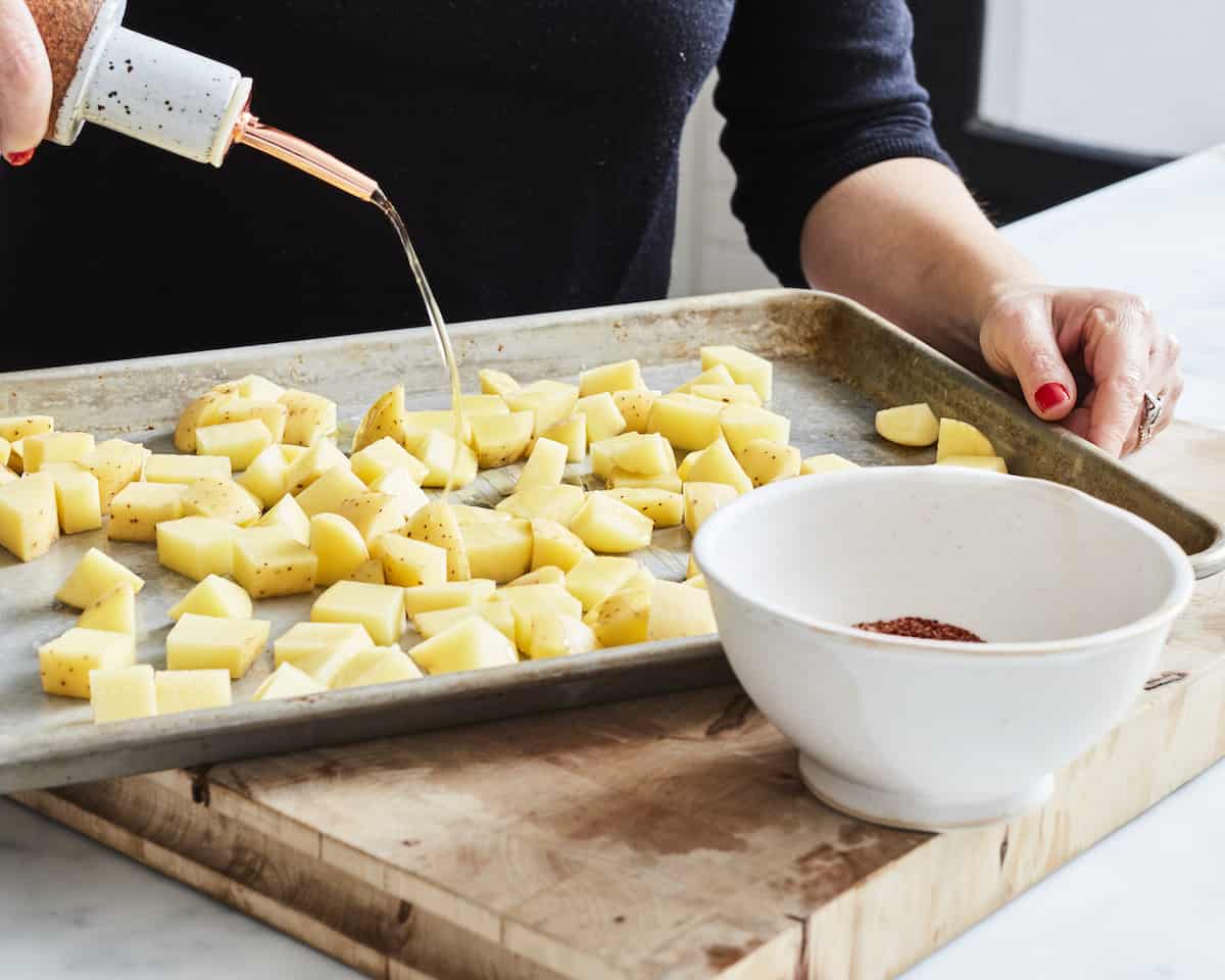 A woman pouring oil through a spout onto a baking sheet full of cubed potatoes with a small white bowl on the side with seasonings.