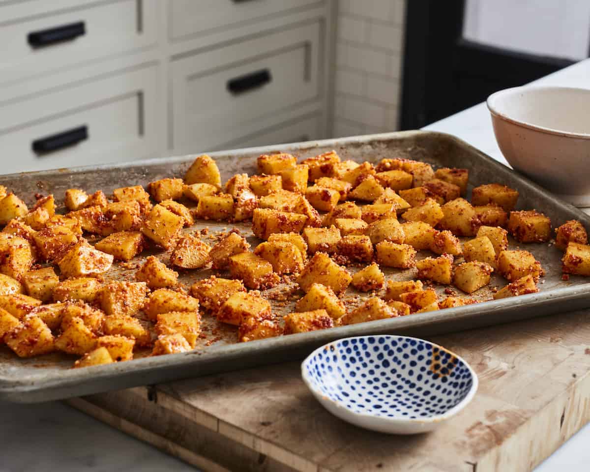 A baking sheet with cubed potatoes, seasoned and coated in grated parmesan, with two empty bowls on the side.