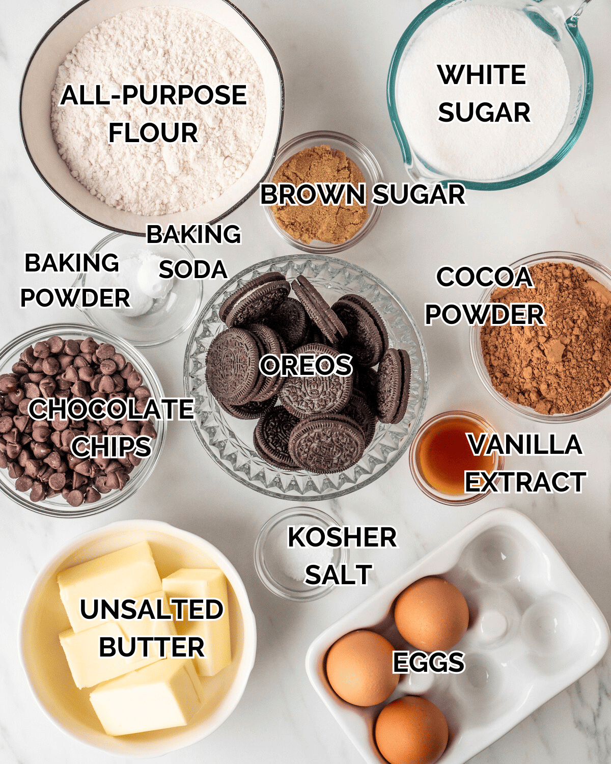 A variety of bowls with all the different ingredients needed to make slutty brownies.  All purpose flour, white sugar, brown sugar, baking soda, baking powder, chocolate chips, oreos, cocoa powder, unsalted butter, kosher salt, vanilla extract, and eggs.